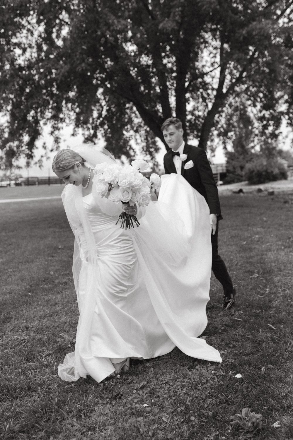 Black and white photo of newlyweds walking outdoors with flowing wedding dress and bouquet.