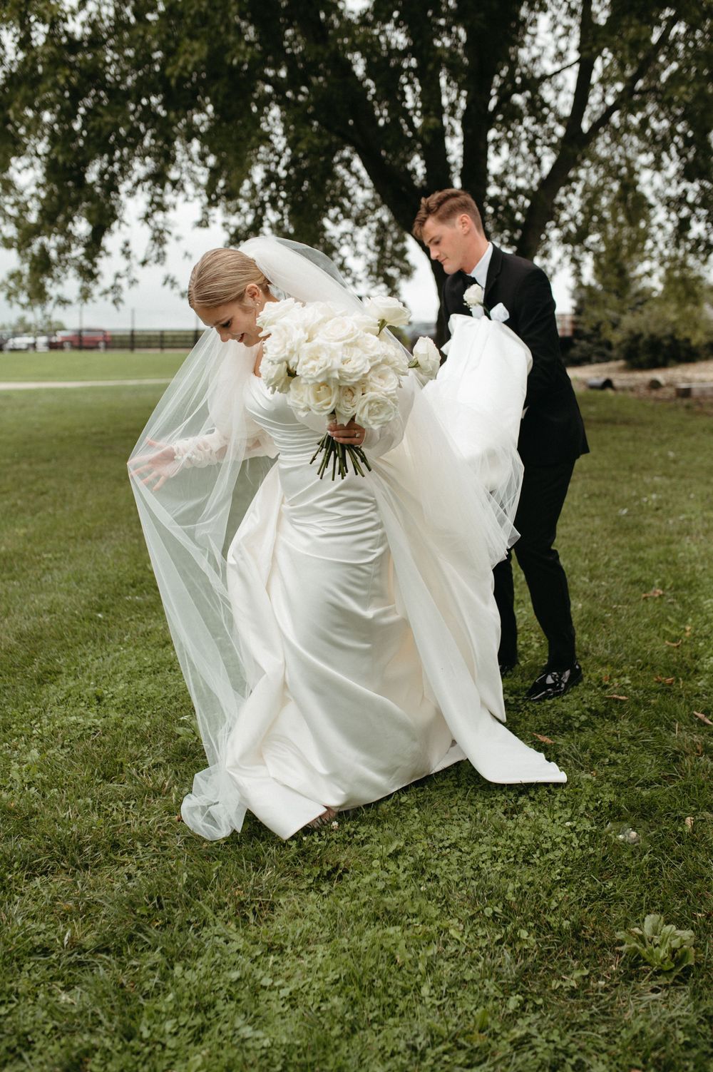 A bride in a white dress and veil holding a bouquet while her dress catches the wind on a grassy lawn.