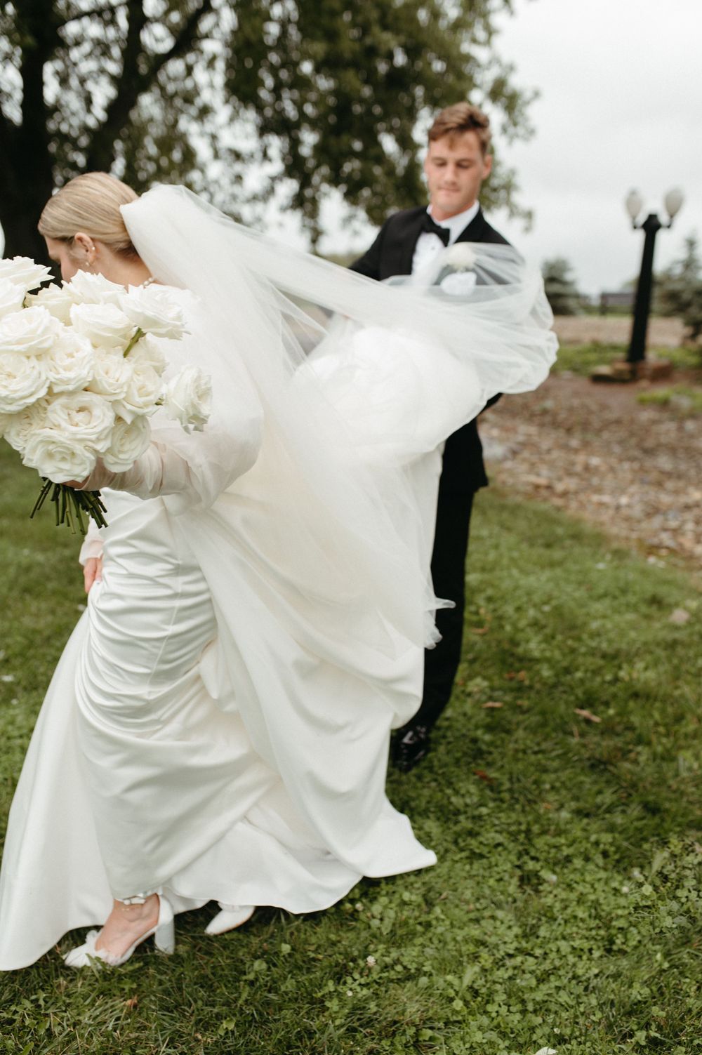 Flowing wedding dress and veil billow in the wind as couple poses outdoors with white roses.
