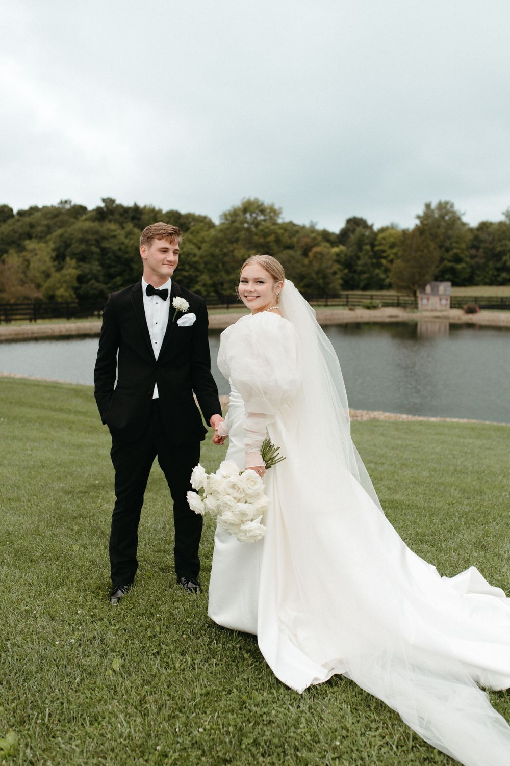 Newlyweds stand together on grassy hill with lake and trees in background.