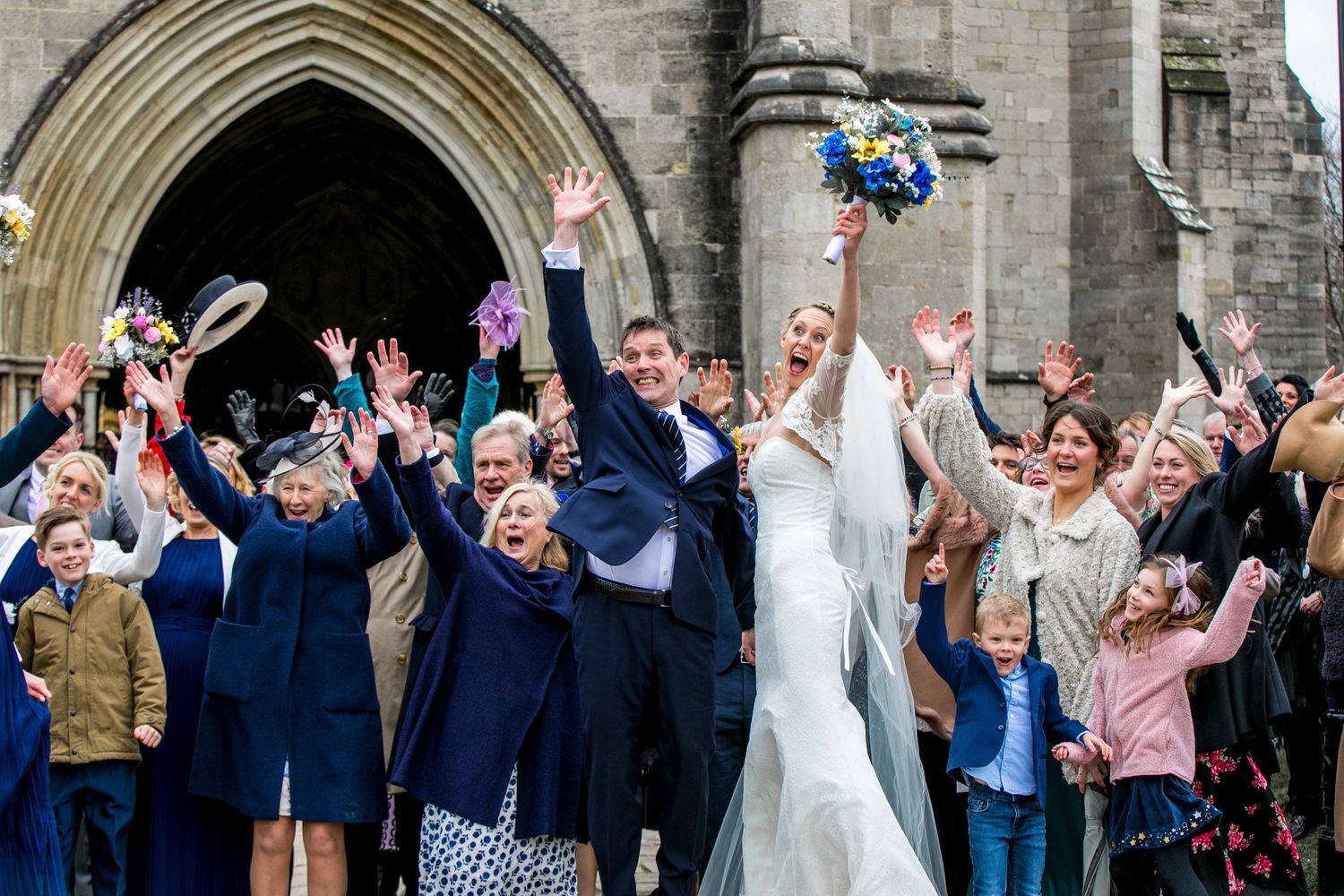 Wedding party and guests celebrate with arms raised outside historic church entrance.