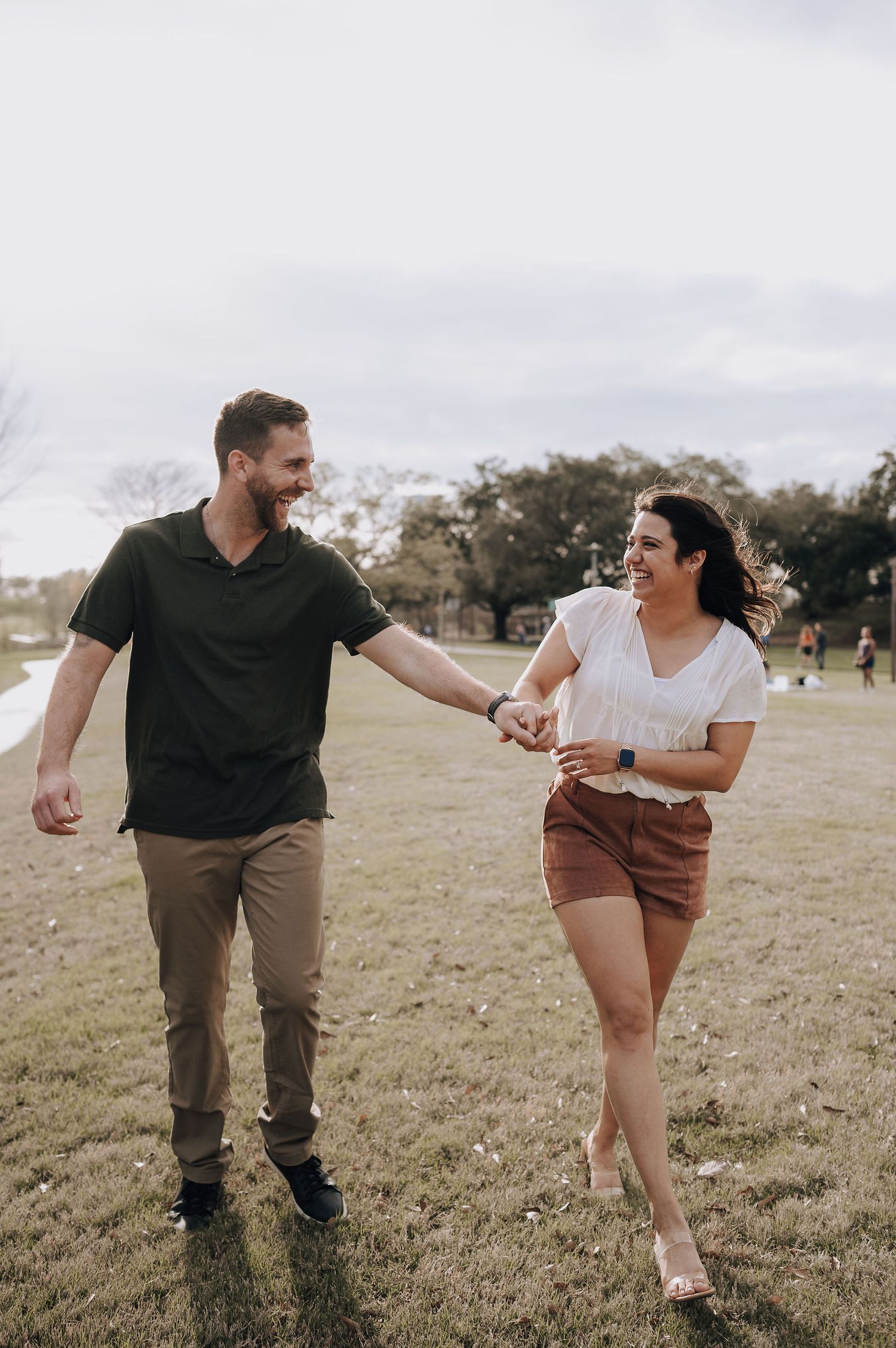 A couple holding hands while walking and laughing together in a grassy outdoor park setting.