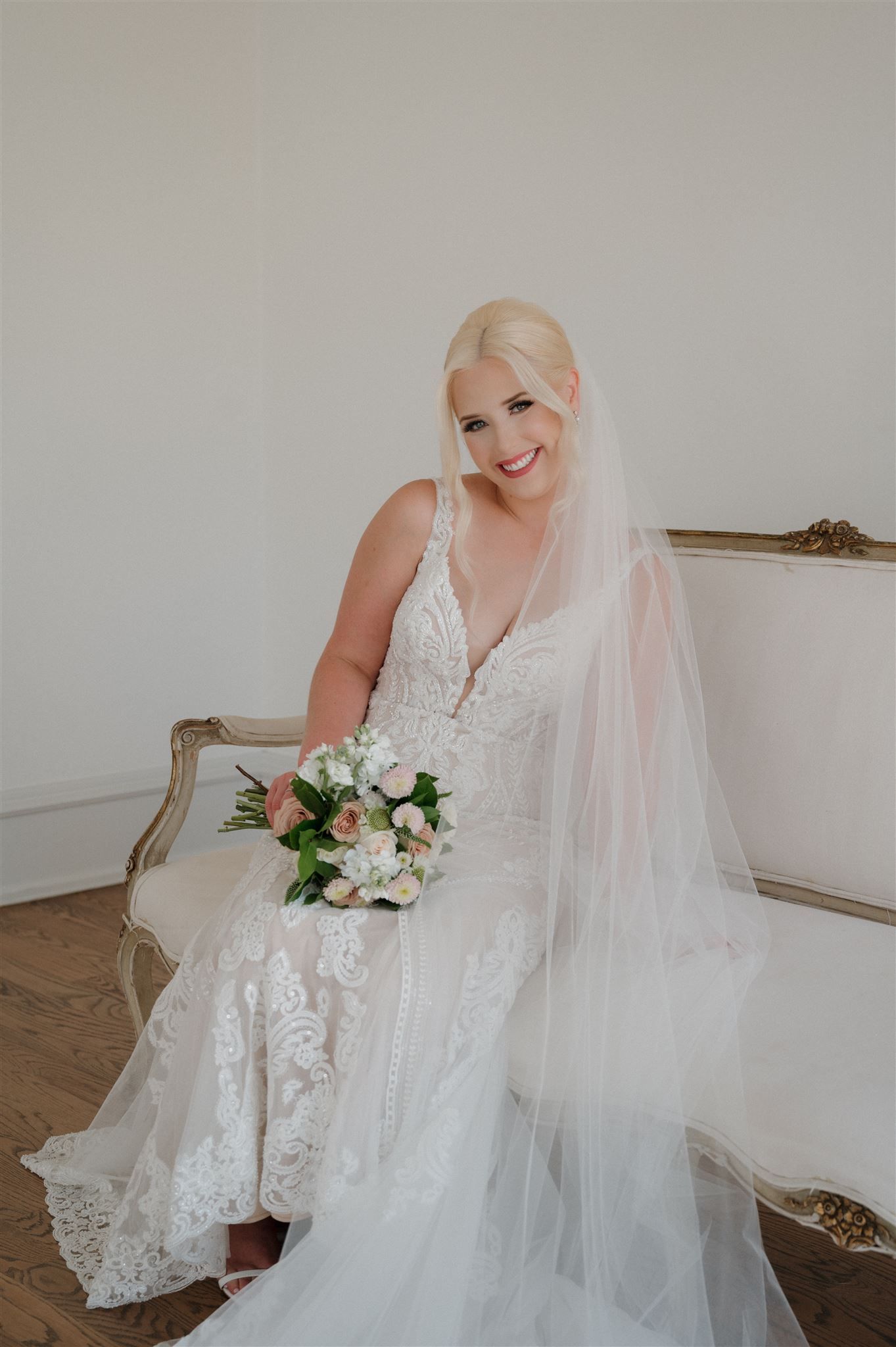 A bride in a detailed lace wedding gown sits elegantly on an antique chair holding a delicate white and pink bouquet.