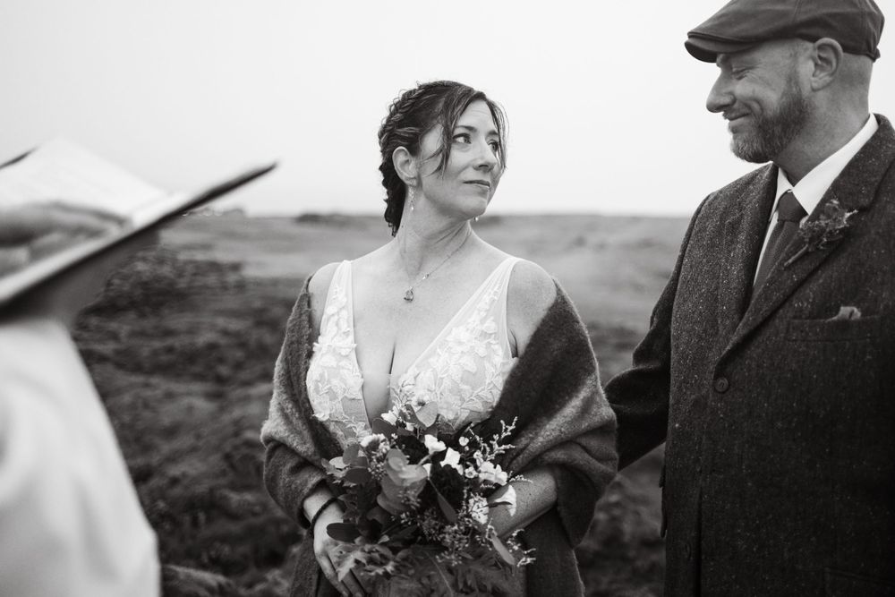 Black and white wedding portrait on windswept coastal cliffs in vintage-inspired attire.