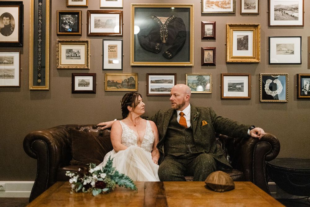 Wedding couple sits on leather sofa surrounded by vintage photographs and artwork.
