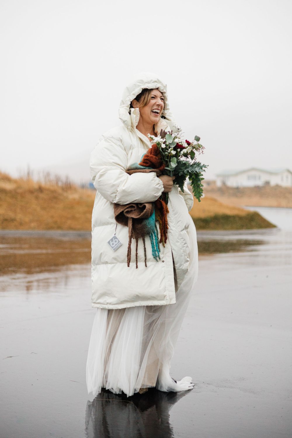 Winter bride in white puffer coat holding bouquet while laughing on beach.