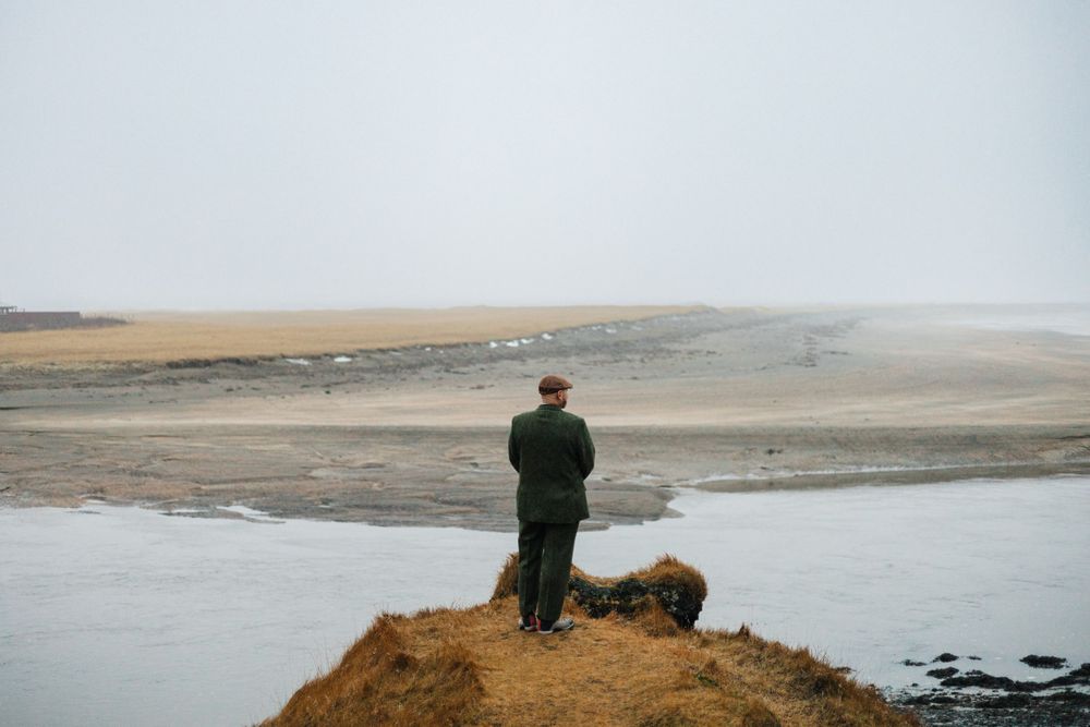 A person in green stands alone on a cliff edge overlooking a misty coastal landscape.