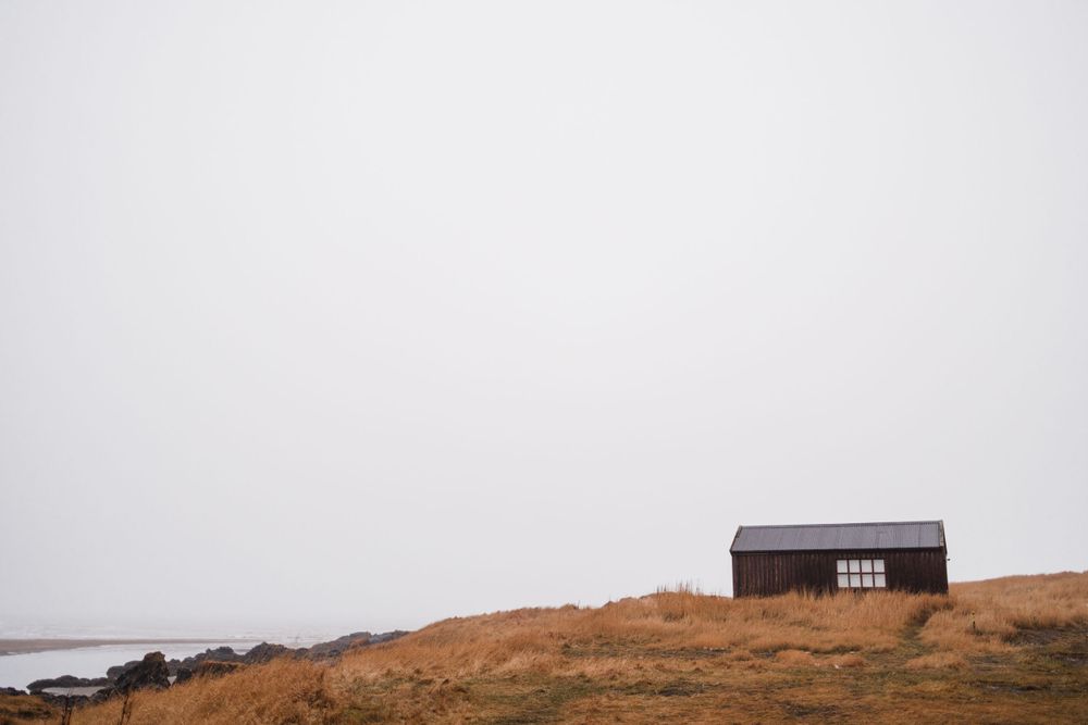 A lone black cabin sits atop a golden grass hill against misty sky.