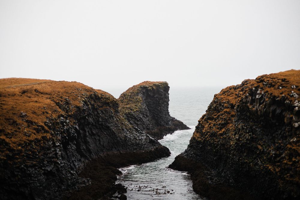 Dramatic black rocky coastal formation with waves crashing between cliffs in moody weather.