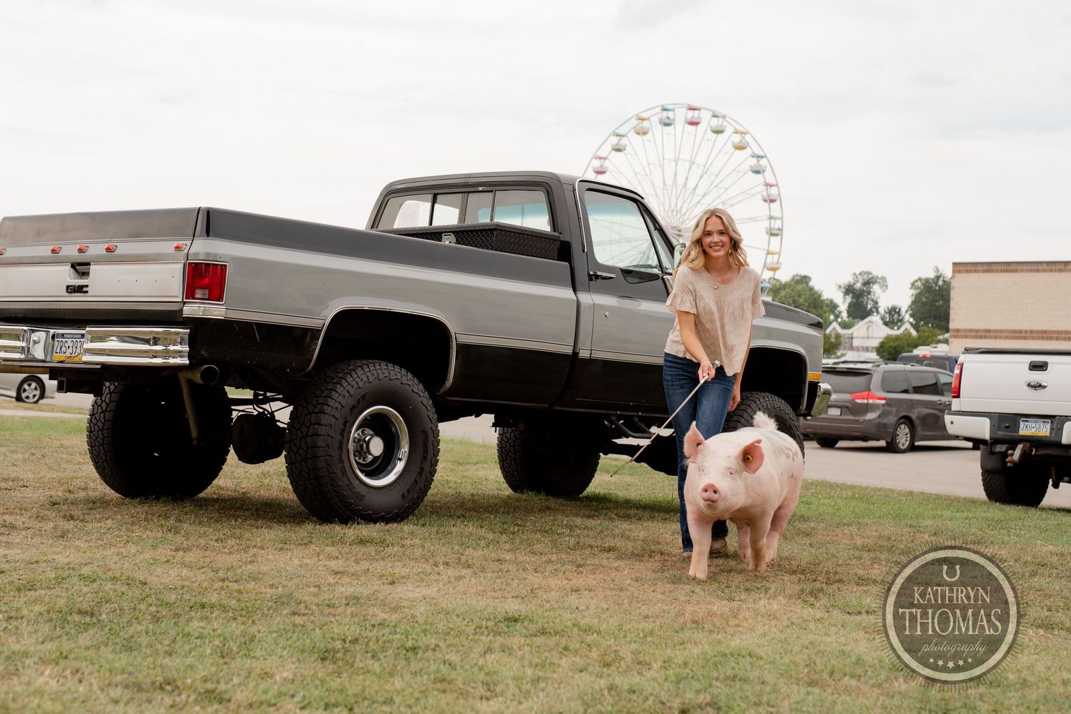 A pink show pig stands by a lifted vintage pickup truck at a county fair with a ferris wheel visible.
