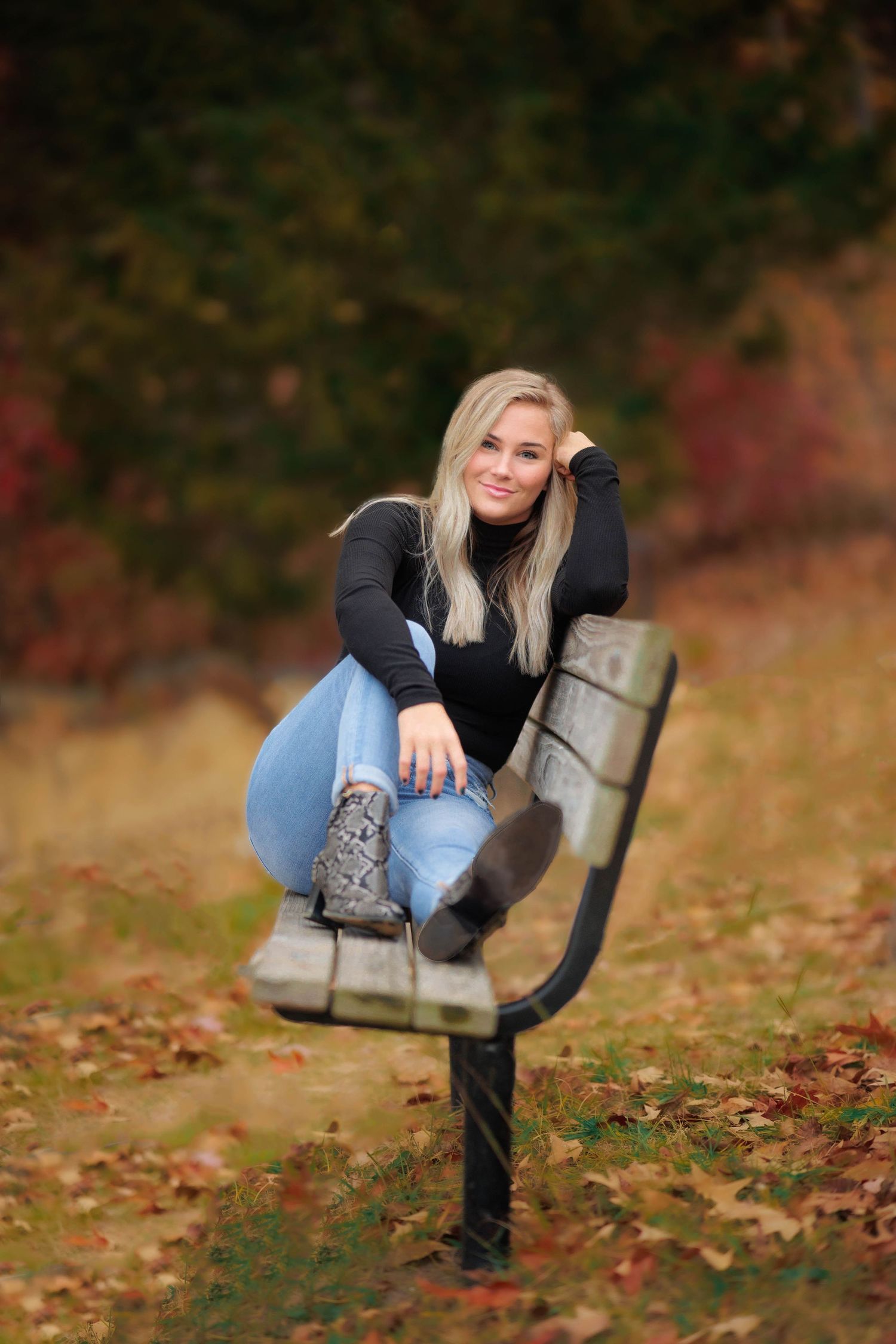 Person in dark sweater and jeans perched on park bench surrounded by autumn leaves.