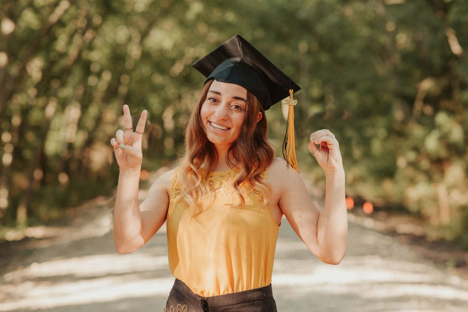 Graduate in yellow top and cap making peace sign on sunny outdoor path during graduation photoshoot.