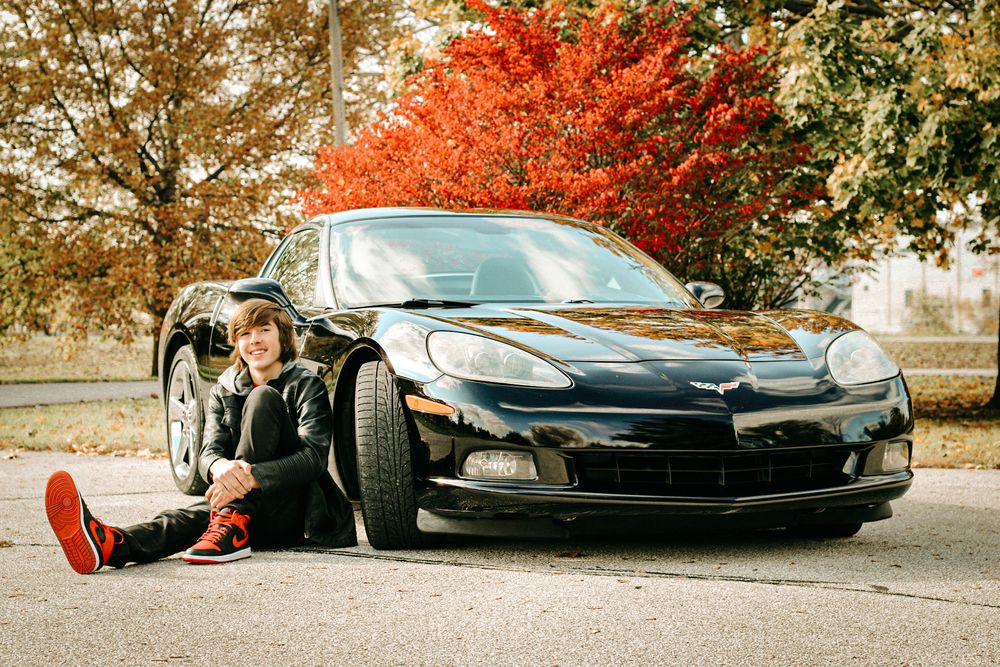 Playful pose on the ground next to a black sports car with red autumn trees in background.
