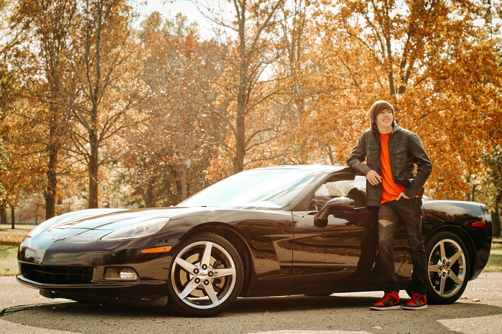 Person in black jacket and orange shirt poses with black Corvette during fall season.
