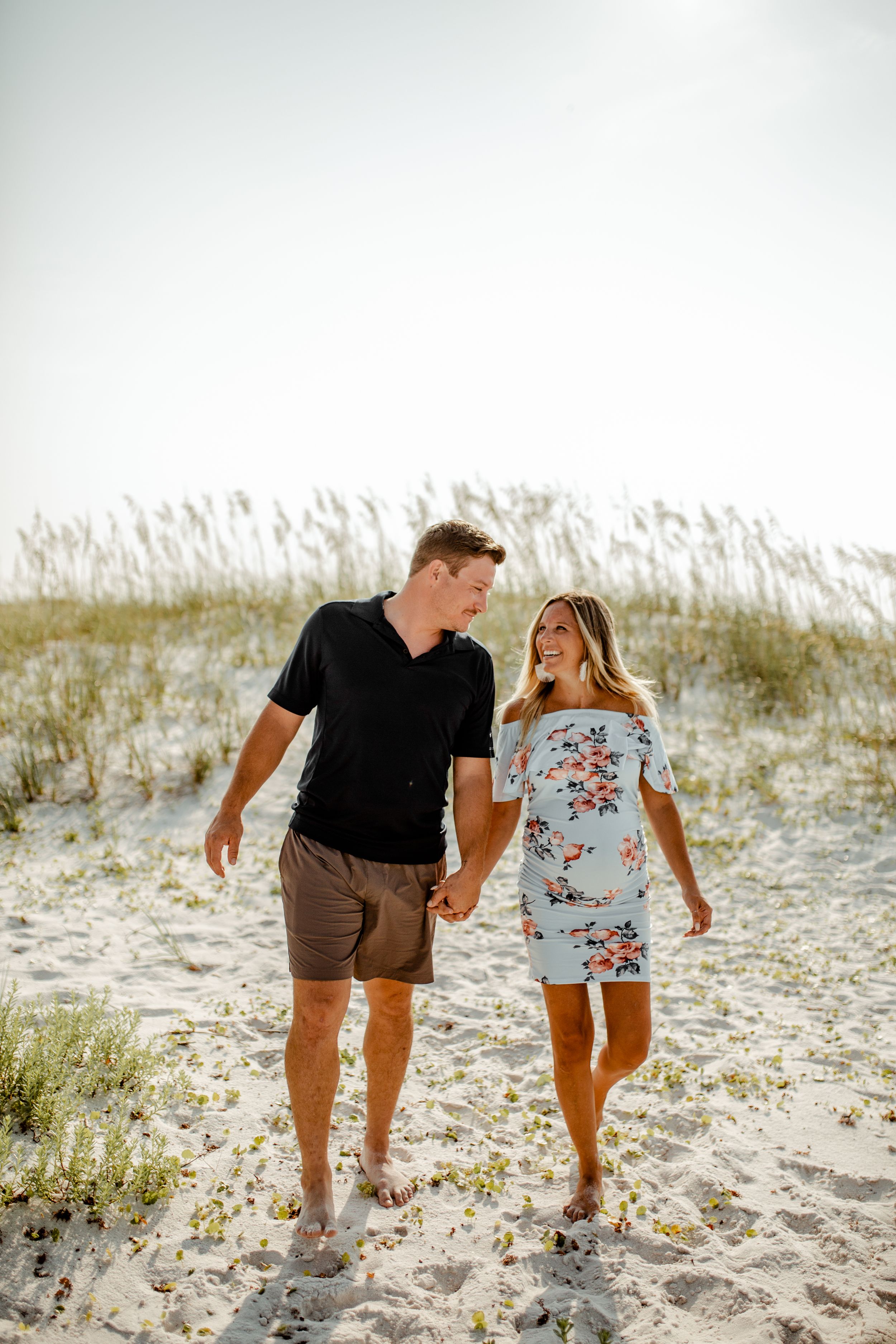 A couple walking hand in hand along a sandy beach path with dunes and grass in the background.