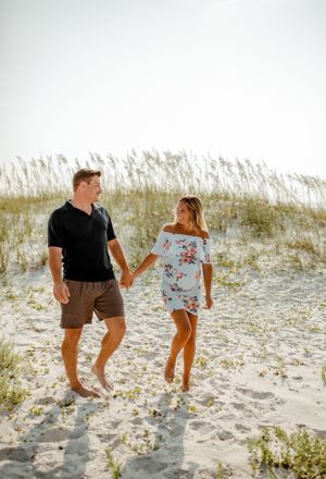 A couple holding hands while walking through sandy dunes with beach grass swaying in the breeze.