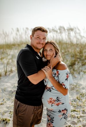 A couple embracing and smiling against a backdrop of coastal dunes and sea grass.