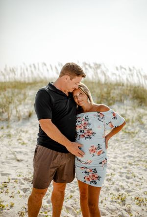 A couple shares a close embrace on sandy dunes with coastal vegetation in soft lighting.
