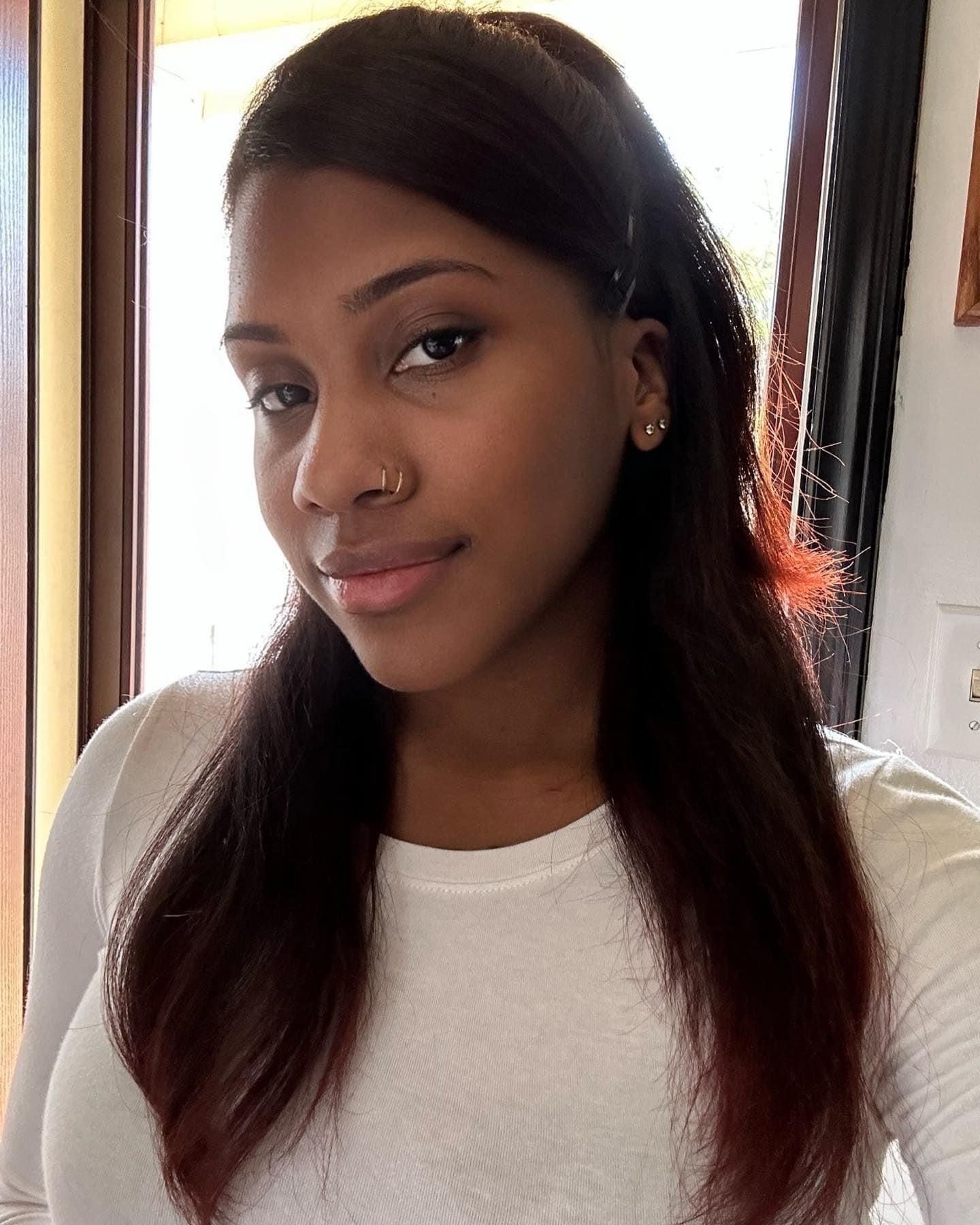 A young Black woman with long dark hair wearing a white shirt takes a casual selfie indoors near a window.