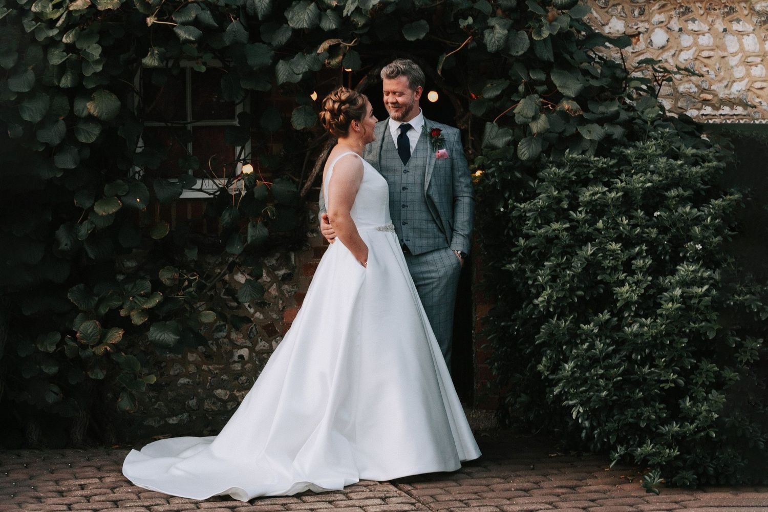 bride in a stunning white dress with a long tail, exchanging a passionate gaze with her groom whilest both hands in her dresses pockets