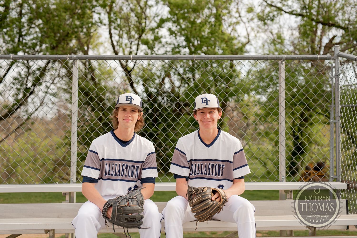 Two baseball players in white uniforms sit together in front of a chain link fence.