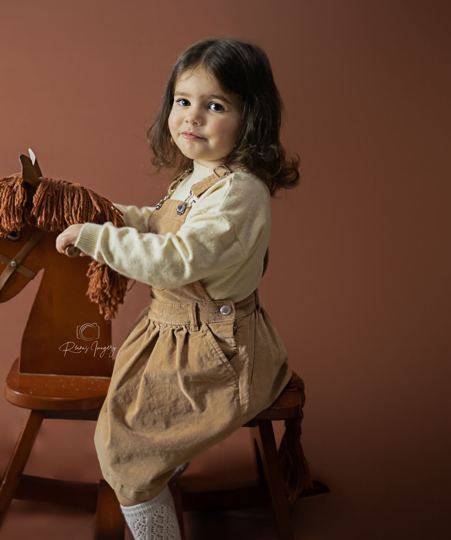 Adorable toddler girl in brown dress posing with a wooden rocking horse in a warm studio portrait.