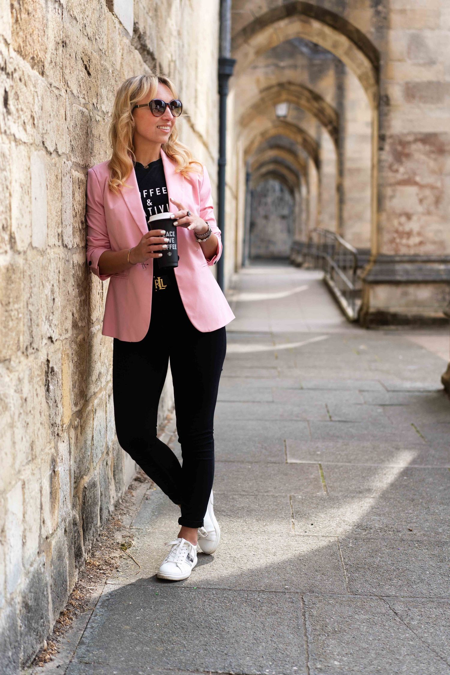 Woman in pink blazer and sunglasses standing in historic archway corridor.