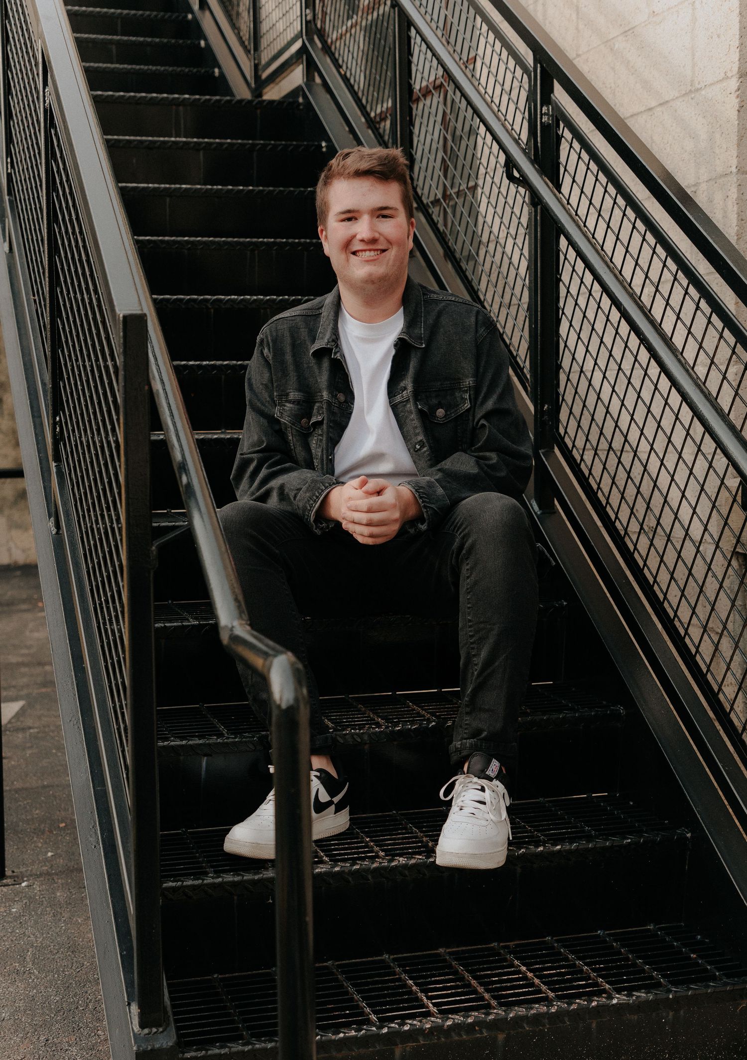 Casual portrait of person sitting on industrial metal staircase wearing denim jacket and white sneakers.