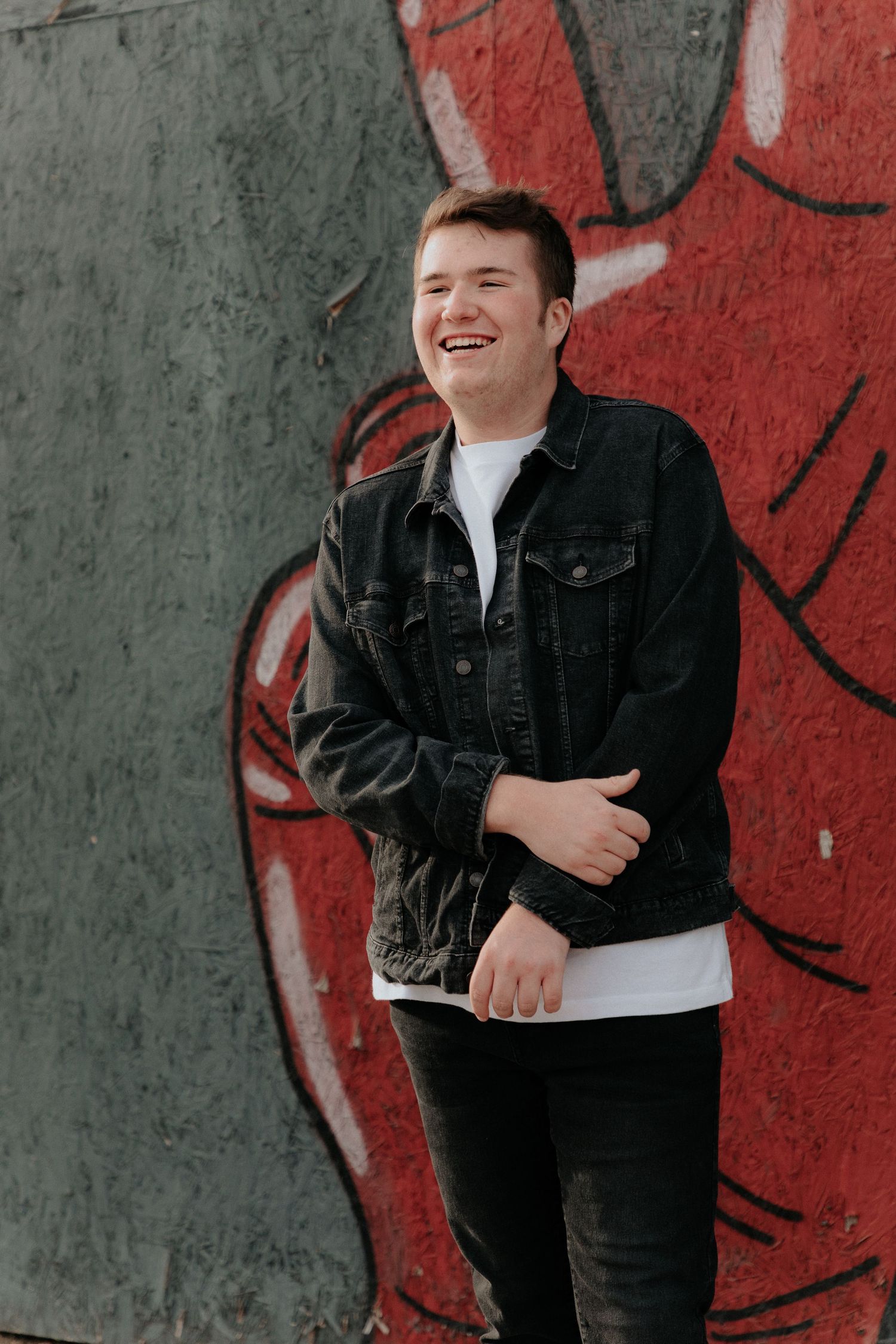 A casual portrait of someone in a black denim jacket laughing against a red and gray graffiti wall.