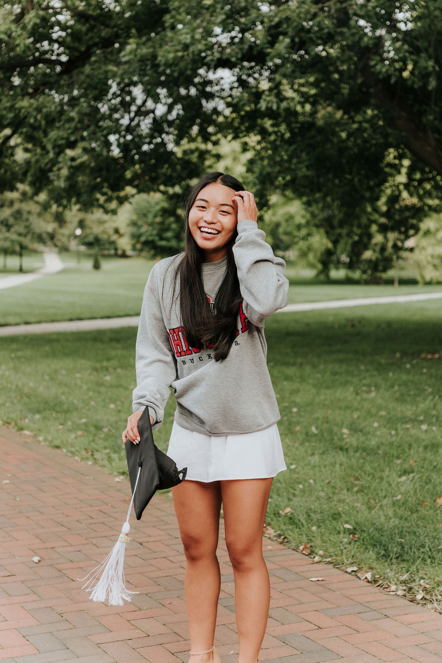 A candid portrait of someone in a gray sweatshirt holding a graduation cap on a brick pathway.