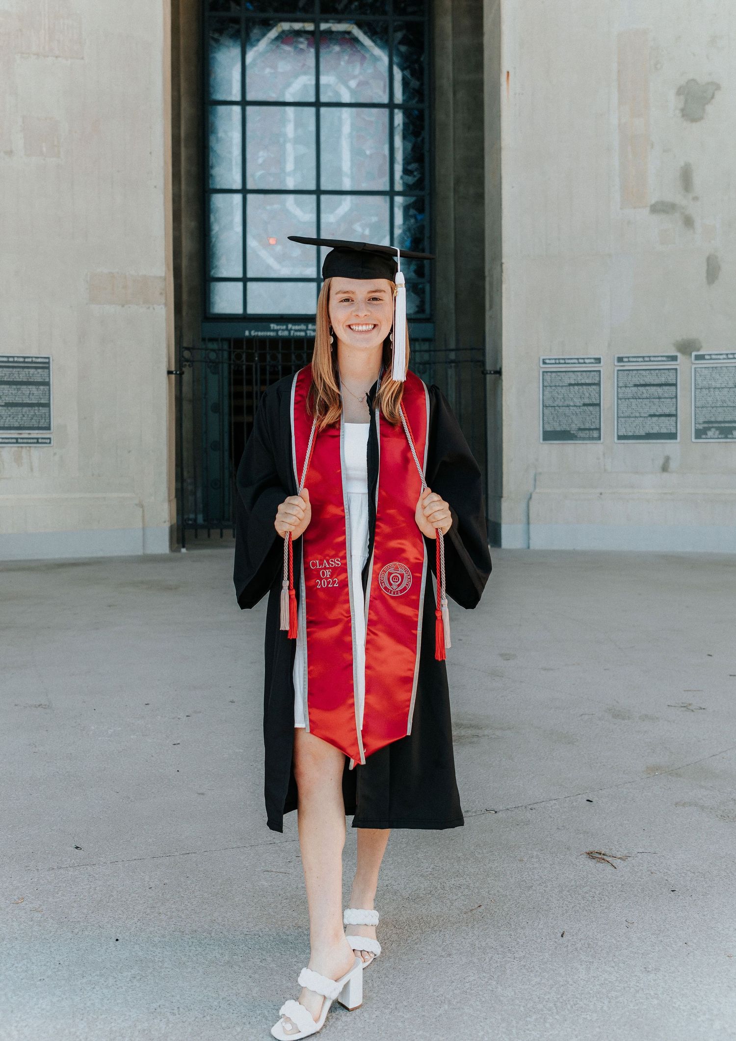A graduate in red and black academic regalia poses in front of an ornate window.