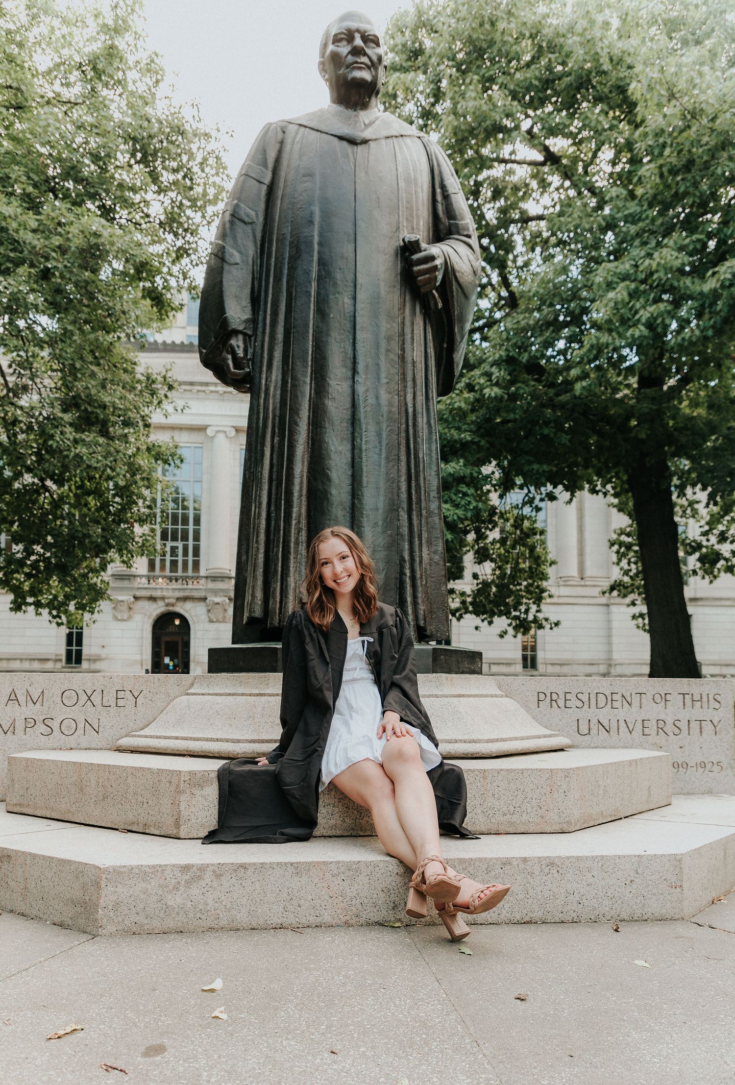 A graduation photo of someone sitting at the base of a large university statue in casual attire.