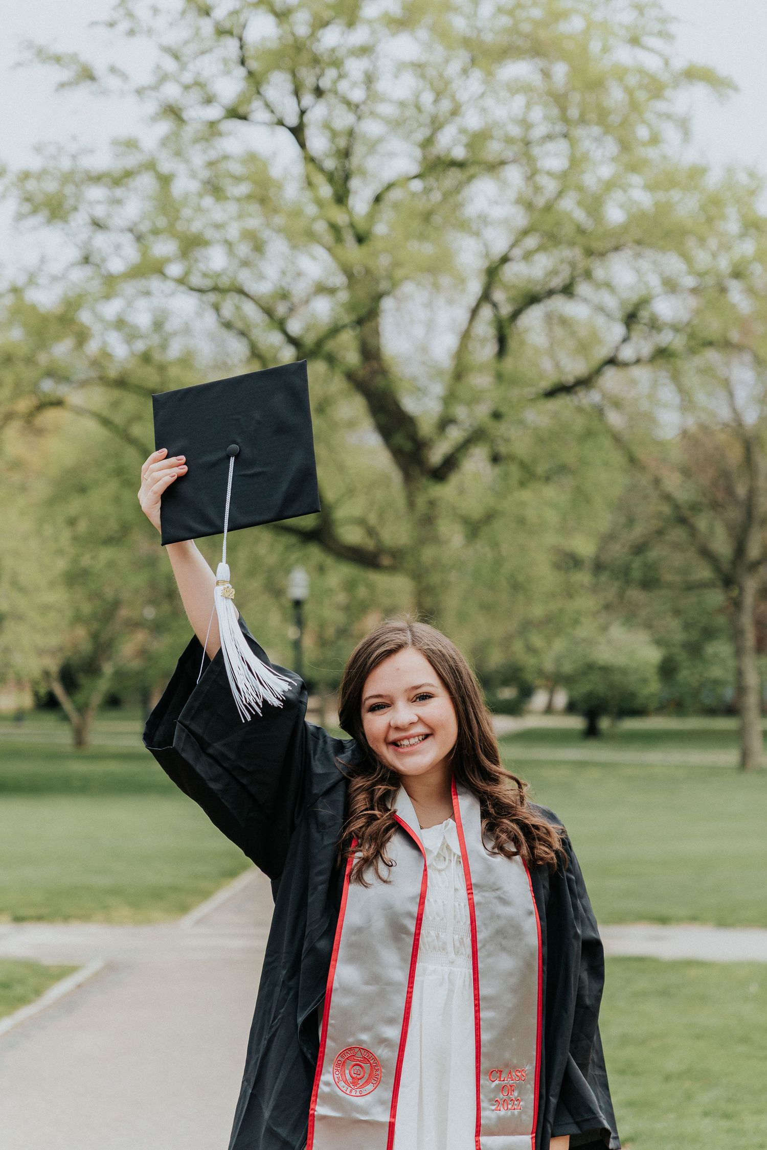 Graduate in black gown celebrates by raising cap in outdoor park setting with trees.