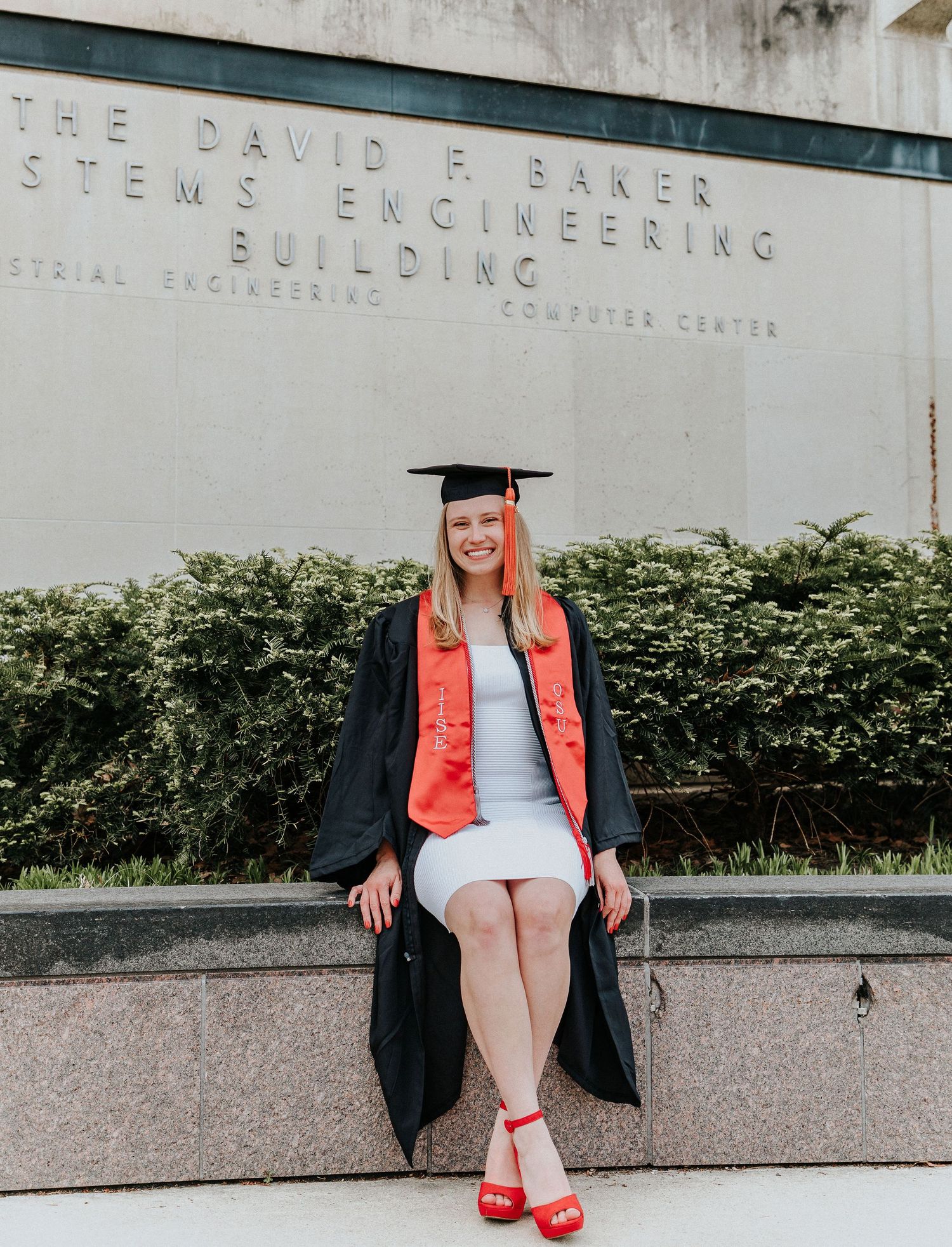 Graduate in red heels poses near engineering building sign wearing cap and gown.
