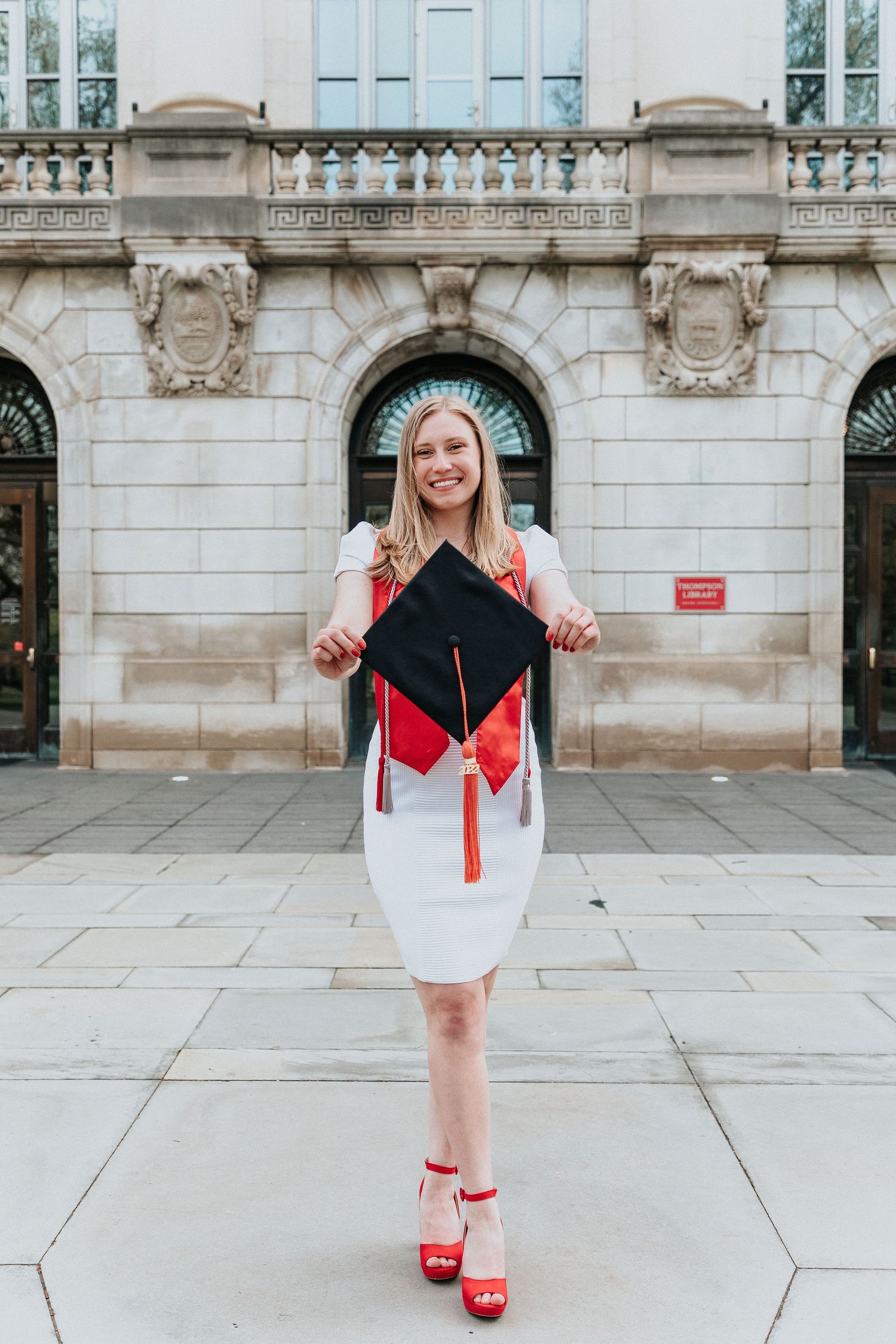 Graduate in white dress holds decorated mortarboard cap while posing in front of ornate stone building.