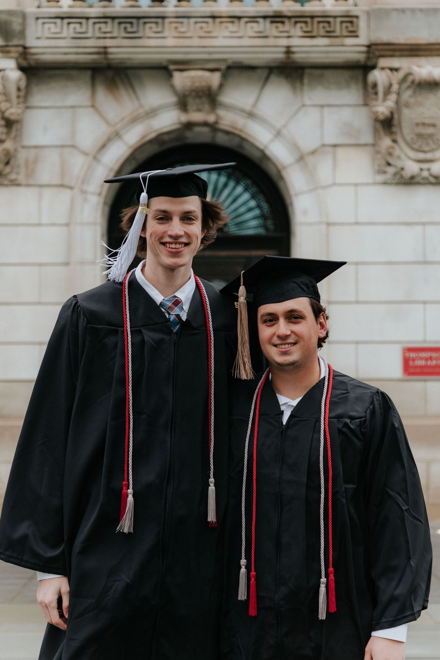 Two graduates in traditional black caps and gowns with red stoles pose in front of stone architecture.