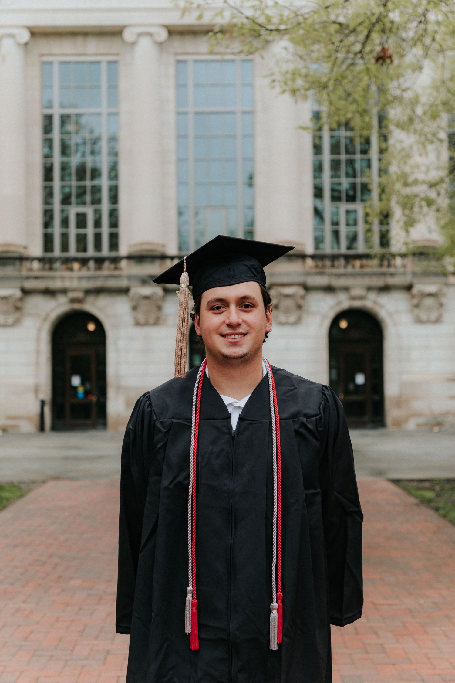 Graduate in black cap and gown smiles while standing in front of ornate university building columns.