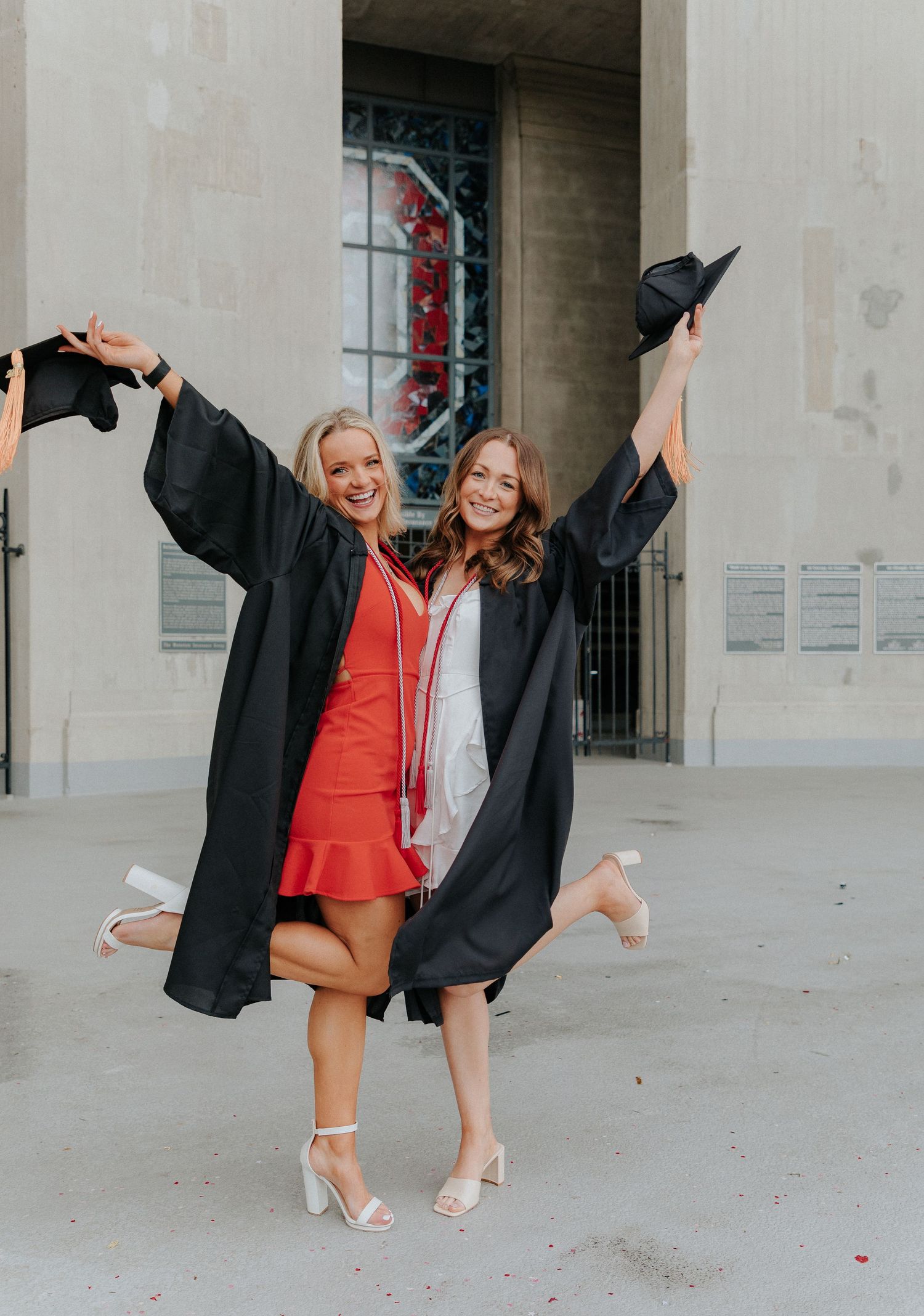 Two graduates in black caps and gowns celebrating with raised arms outside a university building.