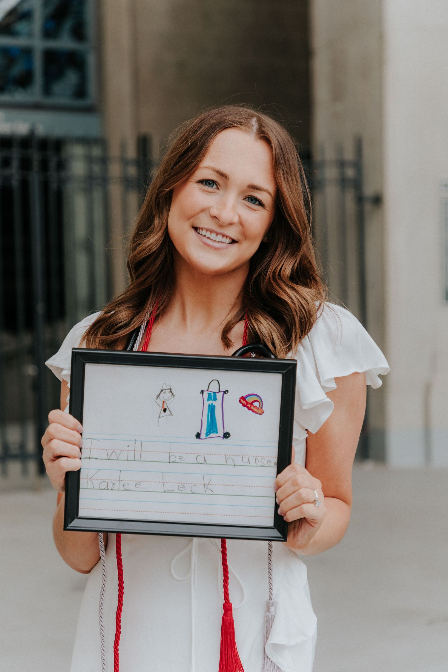 A graduate holding a framed children's drawing while wearing a white dress with red accents.