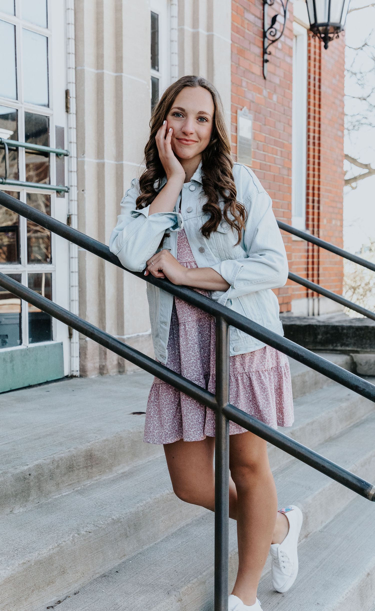 A casual spring outfit featuring a light denim jacket and floral dress paired with white sneakers on stone steps.