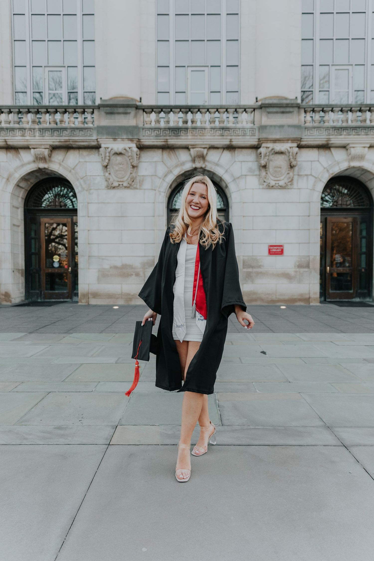 A graduation celebration pose in black cap and gown with a silver dress outside a classic building.