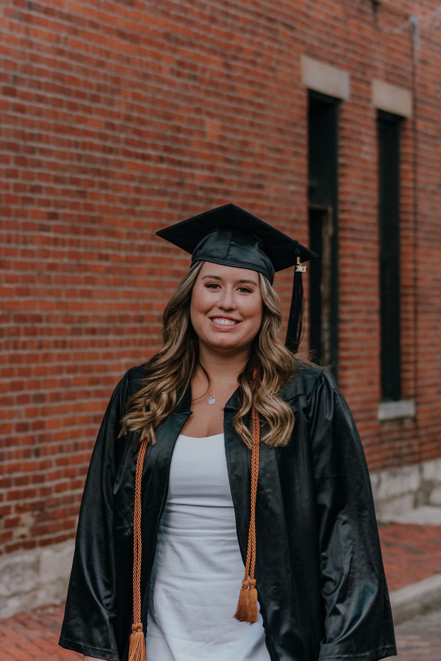 Graduate in black cap and gown smiles in front of brick building wall.