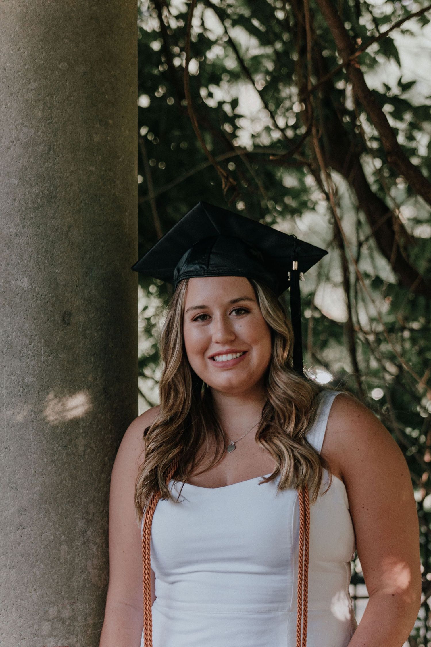 Graduate in cap poses against tree trunk with dappled sunlight in background.