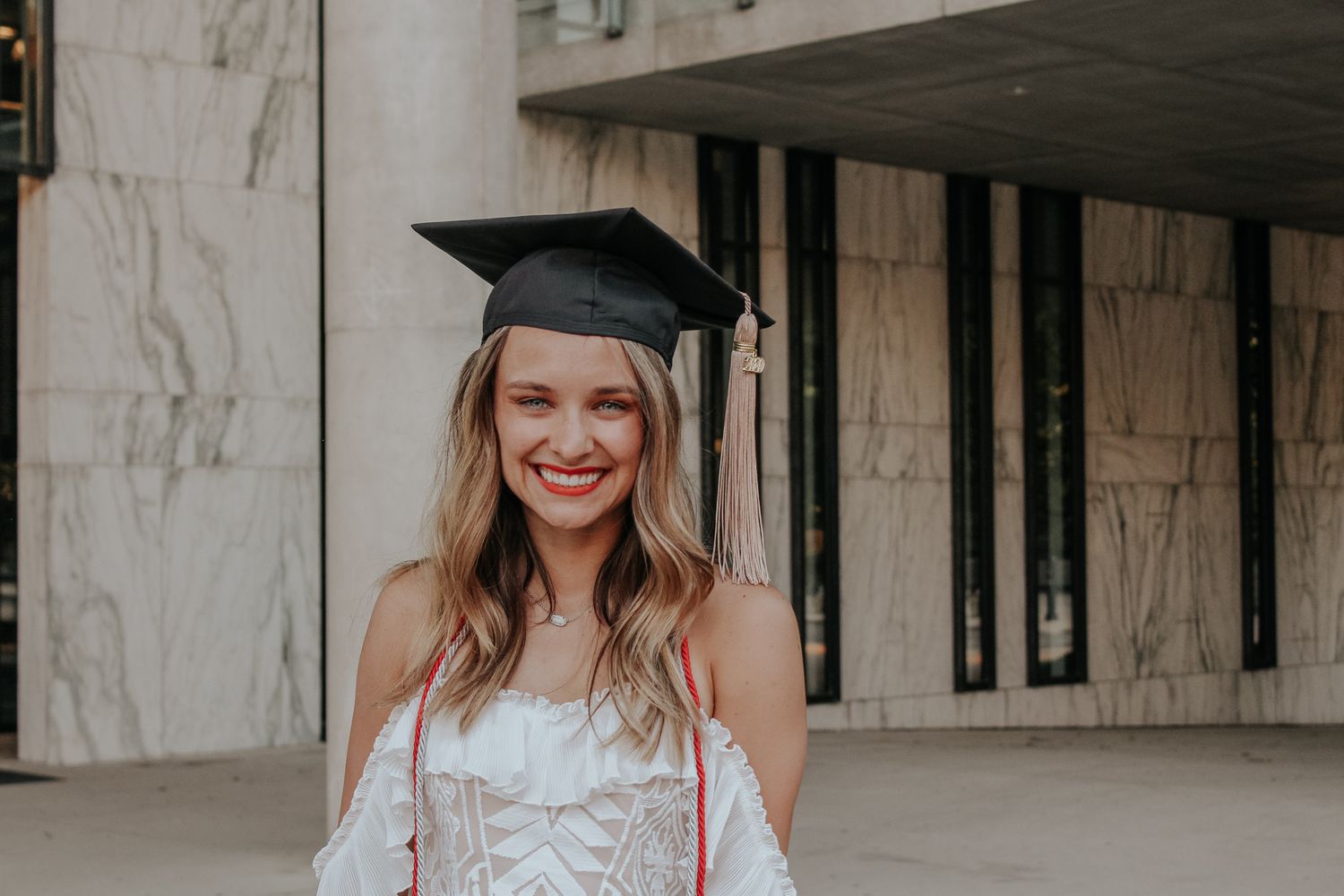 A graduate poses in cap and gown with a bright smile outside a building.