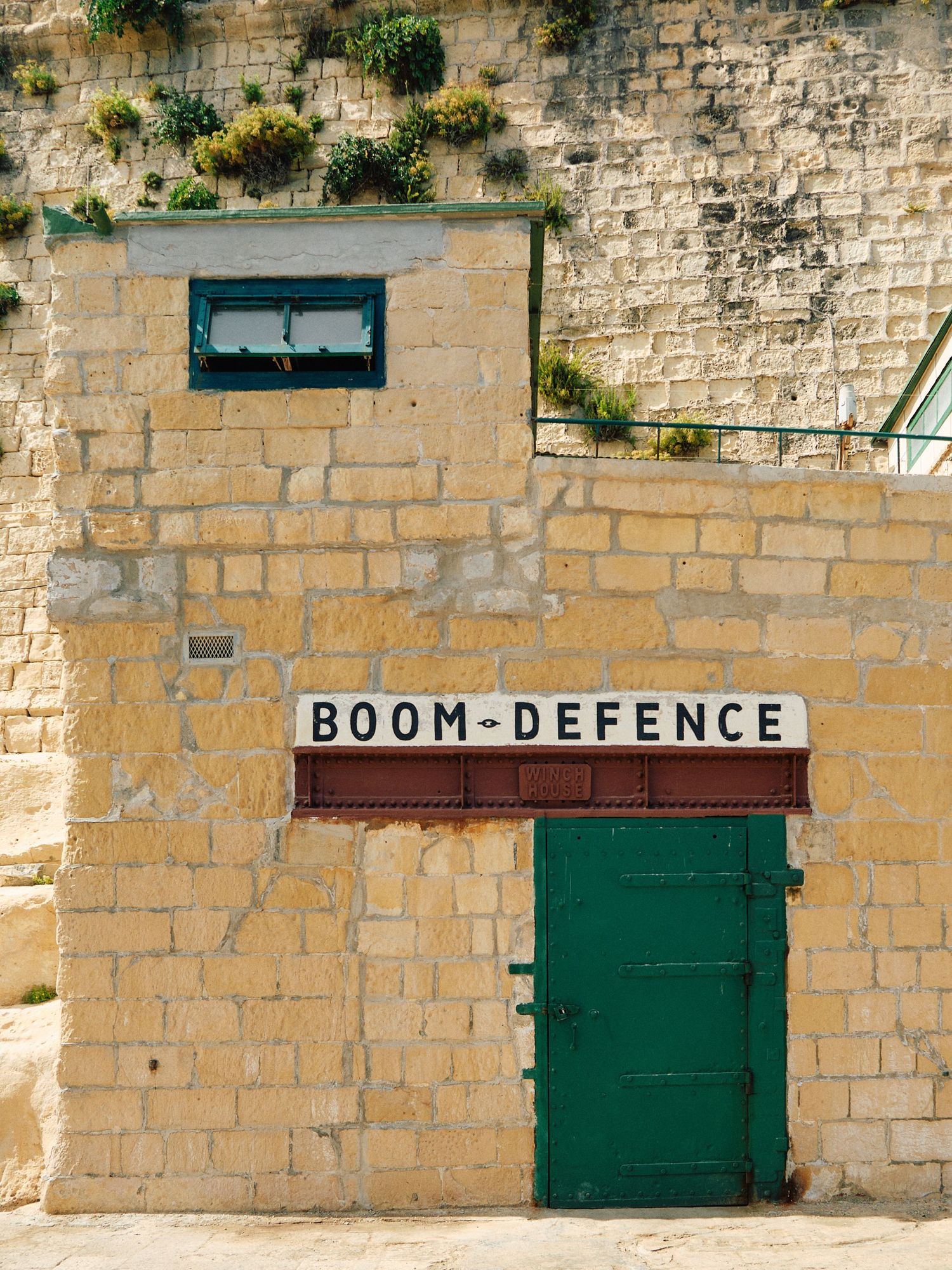 Limestone wall with green door marked 'Boom-Defence' and plants growing from stone.