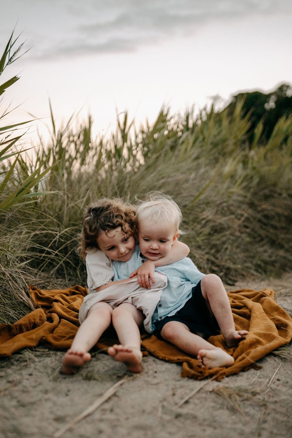 Two children sit together on an orange blanket among tall beach grass.