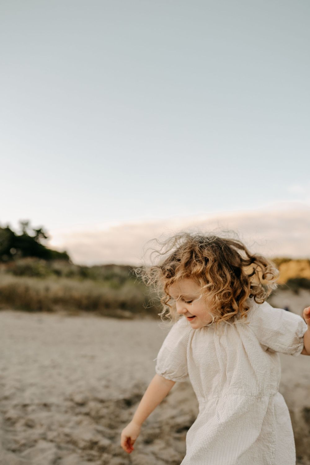Young child with curly hair twirling in white dress on beach at sunset.