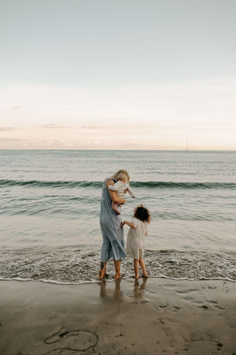 Mother and children standing at ocean's edge during sunset with waves rolling in.