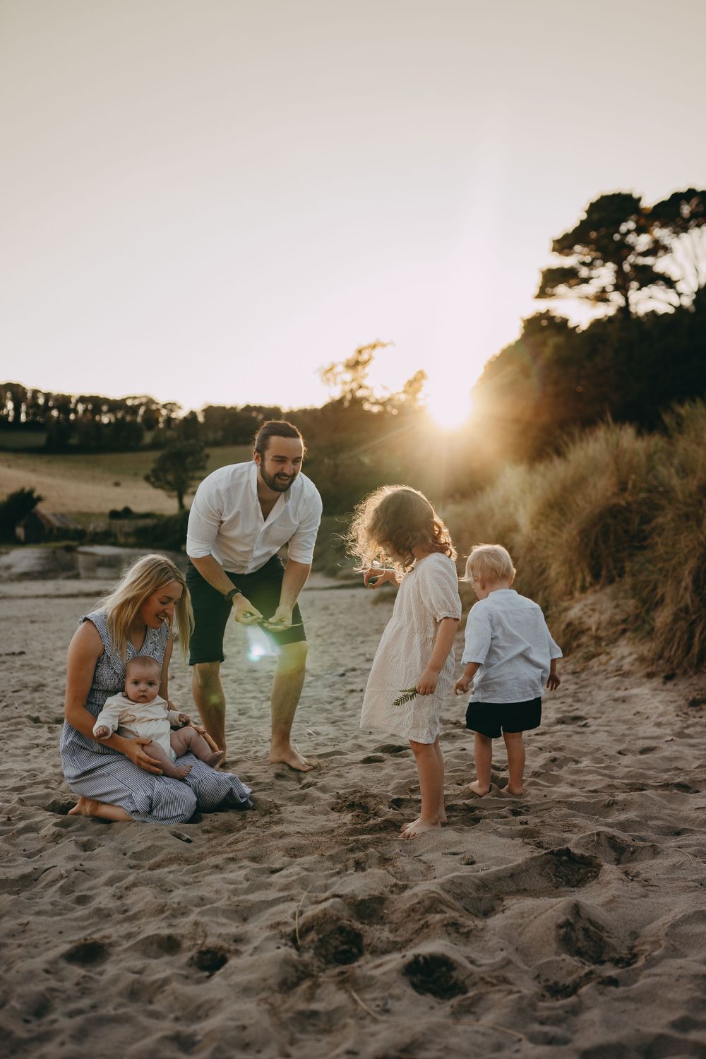 Family gathered on sandy beach during golden hour with sun setting behind coastal trees.