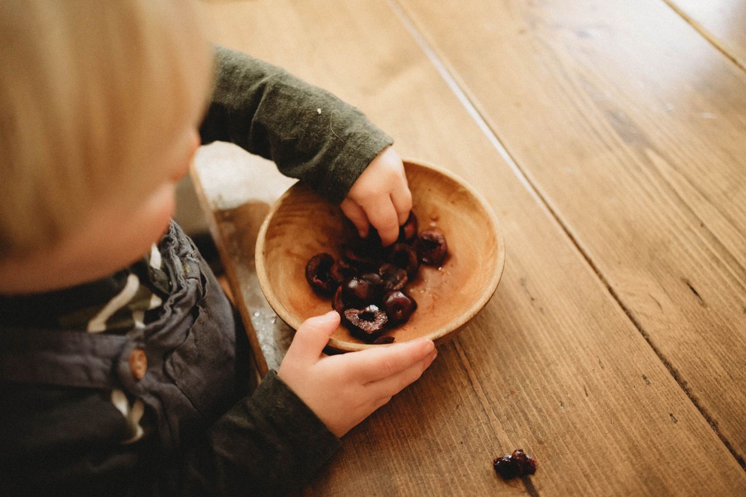 Child's hands holding wooden bowl with dark objects on hardwood floor.